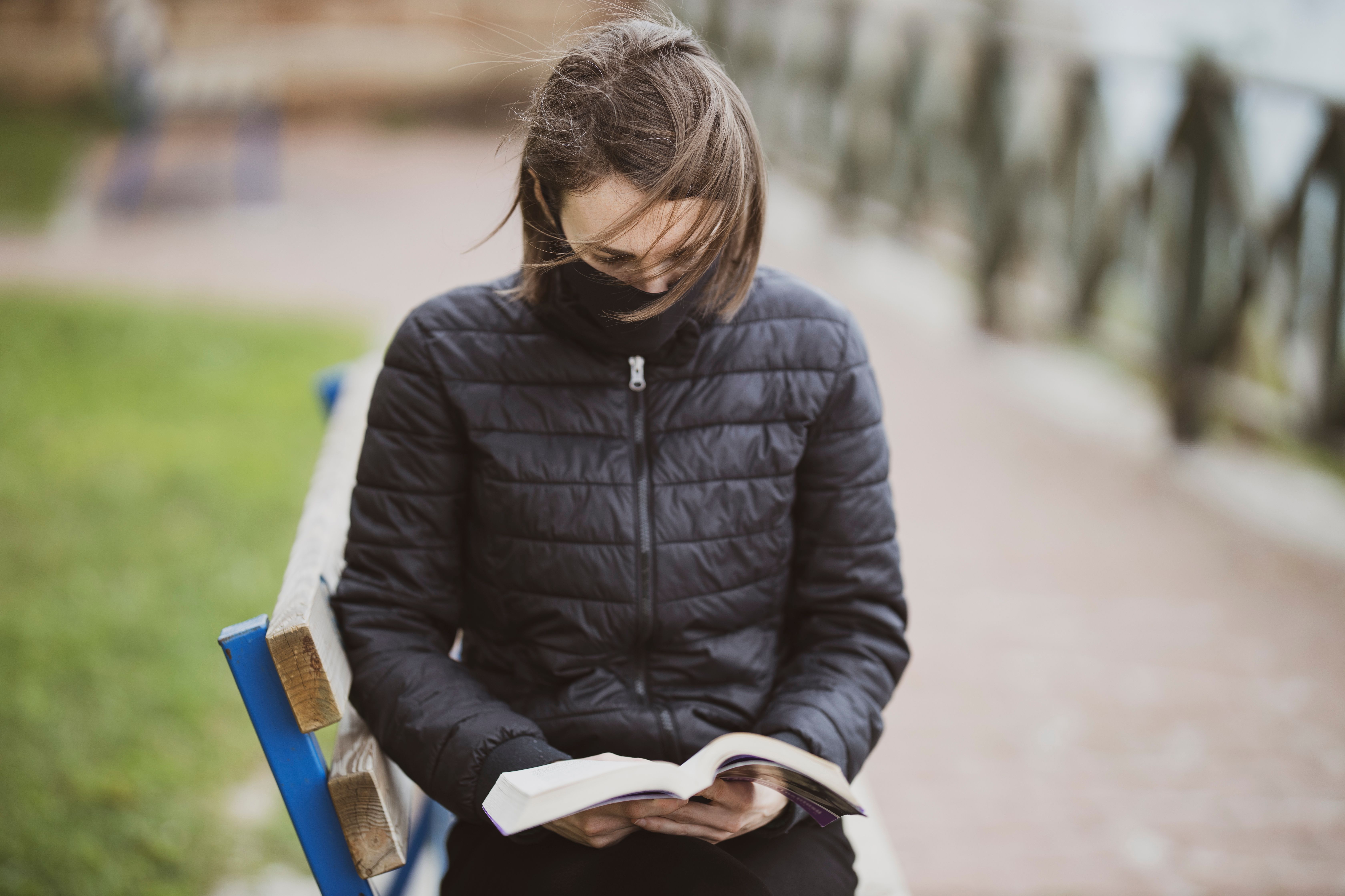 A person sitting on a park bench reading a physical book, with a smartphone face-down on the bench beside them, not being touched
