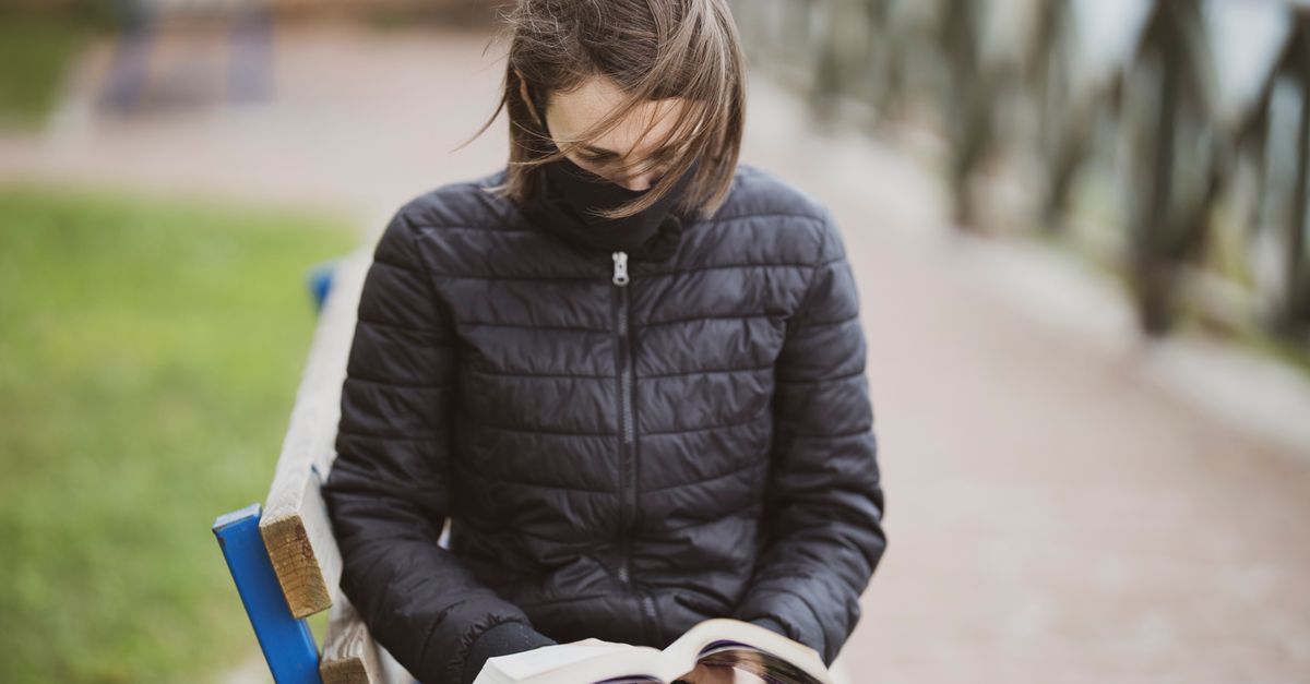 A person sitting on a park bench reading a physical book, with a smartphone face-down on the bench beside them, not being touched