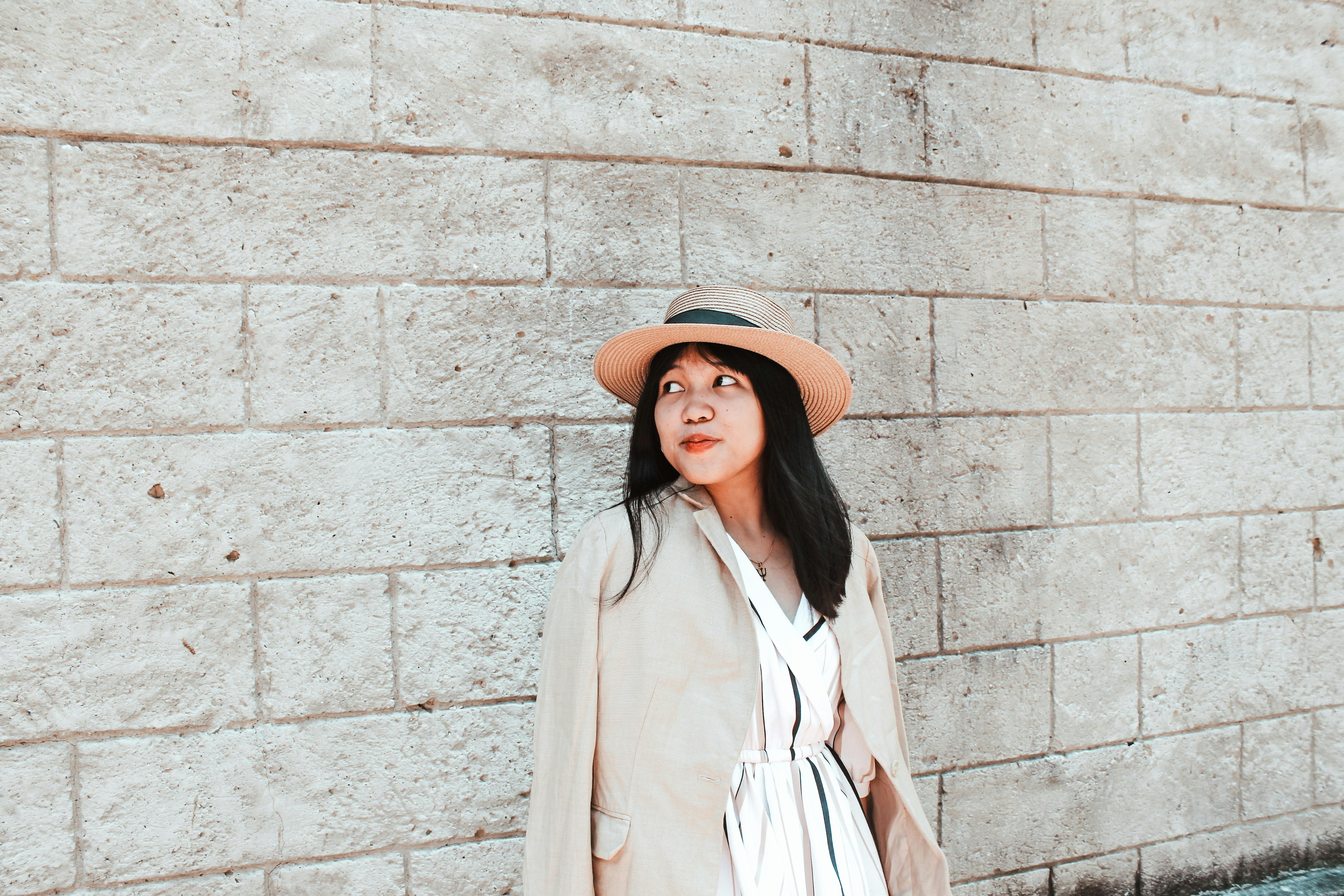 A fashionable woman in a hat and jacket poses against a textured stone wall outdoors.