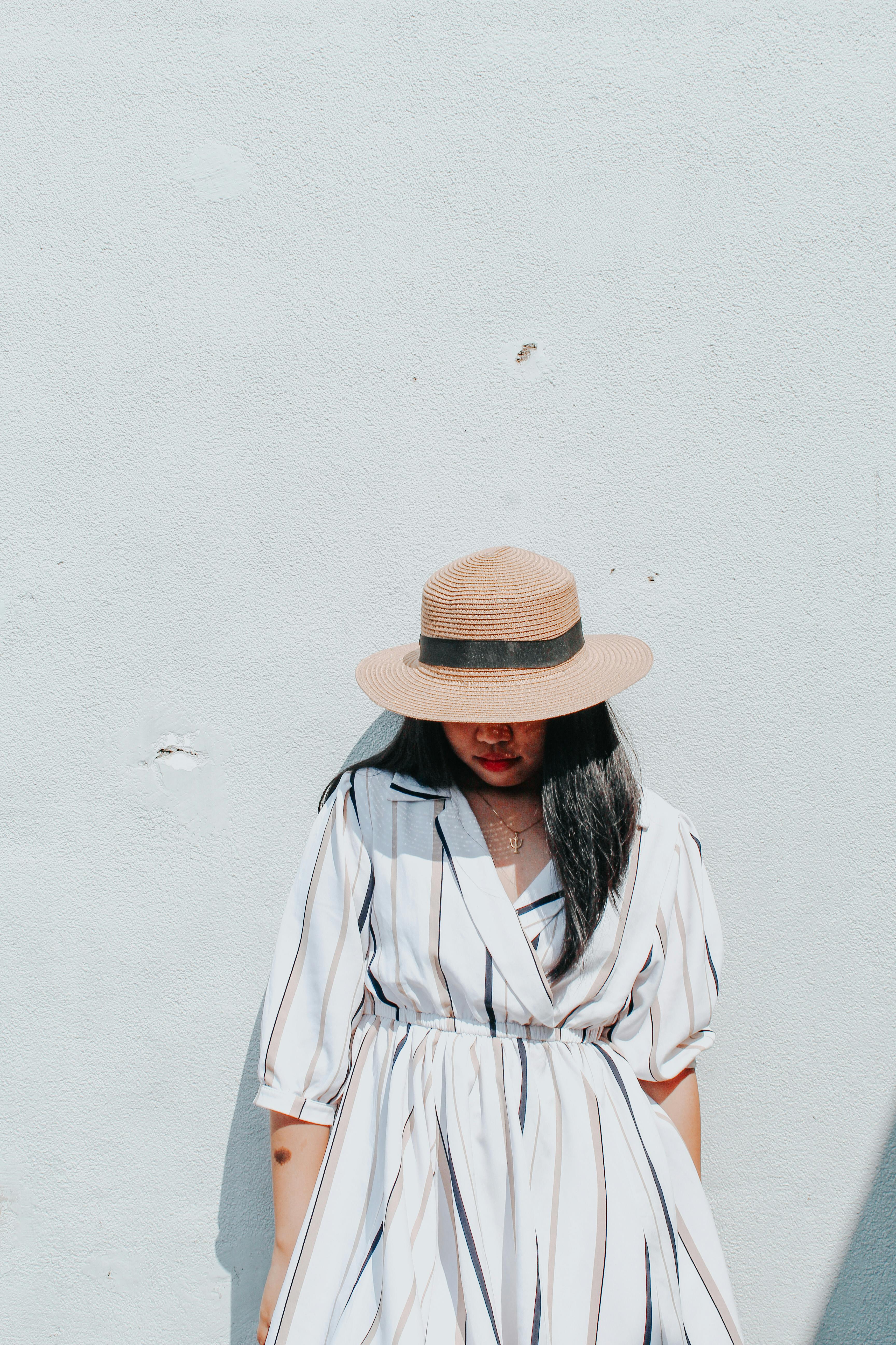 Fashionable woman in a striped dress and hat posing against a minimalist wall, ideal for summer fashion themes.