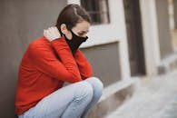 一位戴着口罩、身穿红色毛衣的年轻女子独坐户外，象征着个人防护与孤独