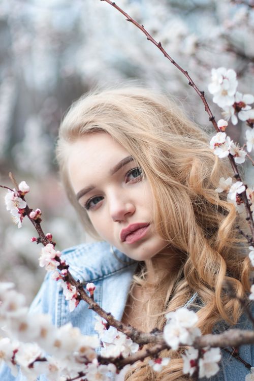 Free Young female with long hair in warm clothes standing near blooming branches of cherry tree and looking at camera in garden on daytime Stock Photo