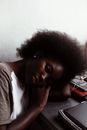 From above of young black woman with curly hair in casual clothes sitting and putting head on table during break on daytime