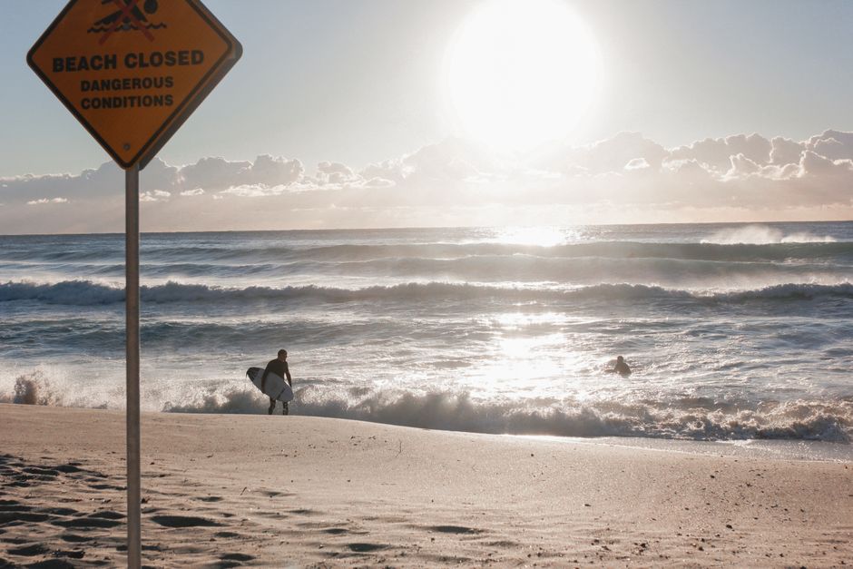 Surfer defies beach closure at Bronte, Australia, with dangerous waves crashing in the sunlight.