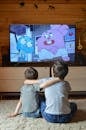 Back view of anonymous adorable little siblings cuddling while sitting on floor and watching funny cartoon on TV at home