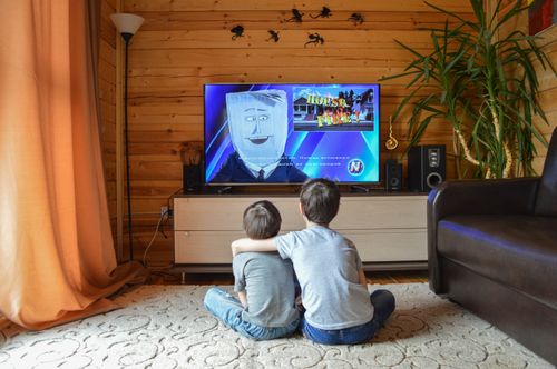 Free Back view of faceless preteen boy in casual clothes hugging little brother while sitting together on floor and watching TV Stock Photo