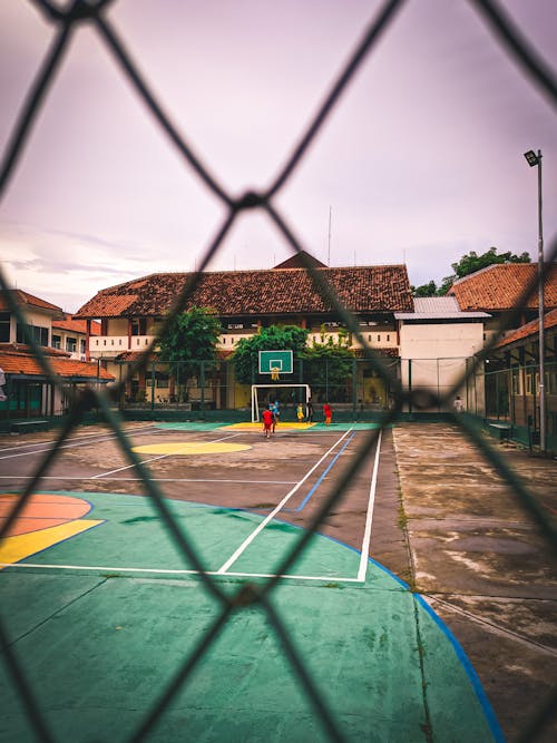 Through metal mesh fence of anonymous people playing basketball on sports ground in town against cloudy sky during sunset