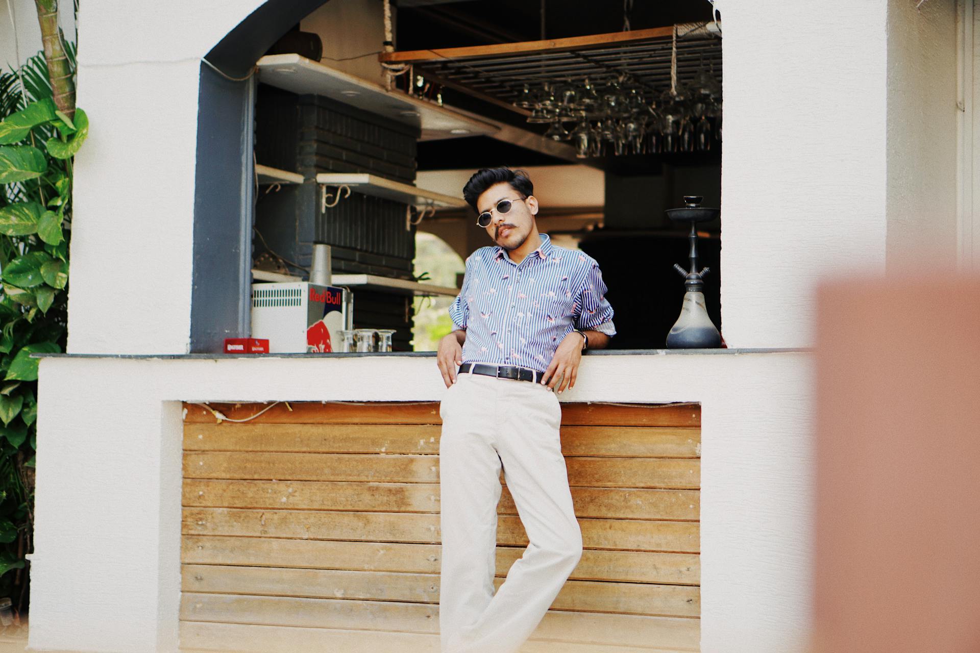 Confident stylish young ethnic male with mustache in casual outfit and sunglasses leaning on counter of outdoor cafe