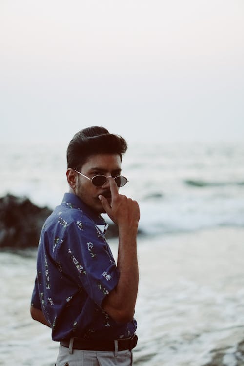 Side view of serious young unshaven Indian guy in trendy outfit adjusting sunglasses and looking at camera while resting near waving sea