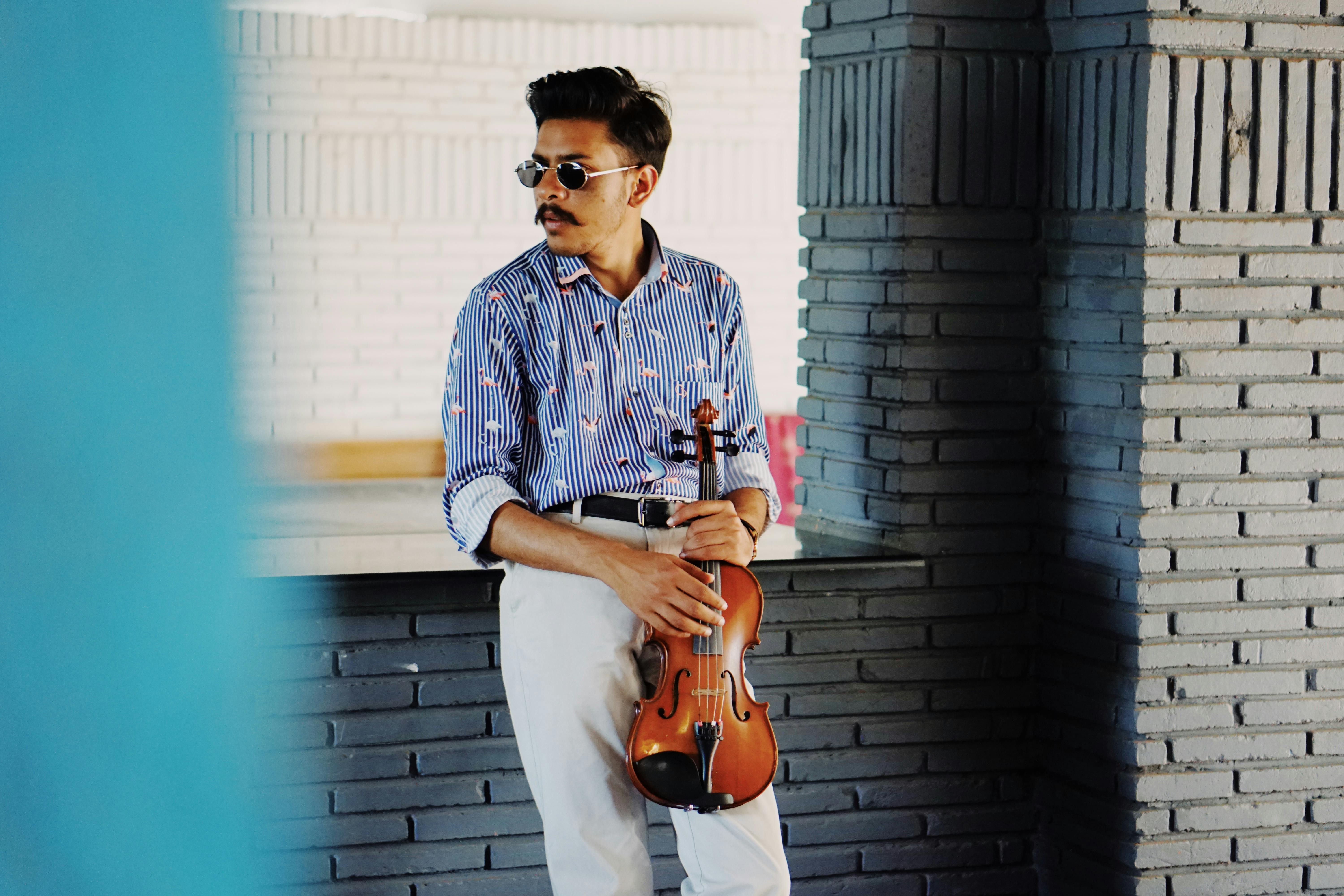Serious young ethnic male musician with mustache in stylish outfit and sunglasses standing in creative studio with violin in hands and looking away
