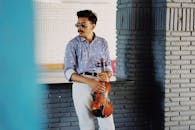 一位表情严肃、留着小胡子、穿着时尚、戴着墨镜的年轻少数族裔男性音乐家，站在创意工作室里，手持小提琴，目光移向别处