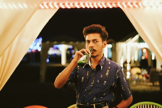 Stylish young ethnic bearded mail in casual outfit drinking bottle of beer and looking at camera while relaxing in outdoor bar at night