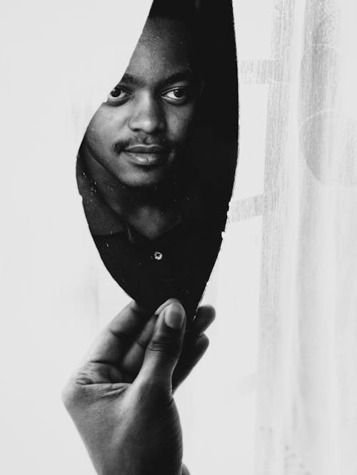 Black and white crop young unshaven ethnic male looking in broken mirror piece standing near window on sunny day