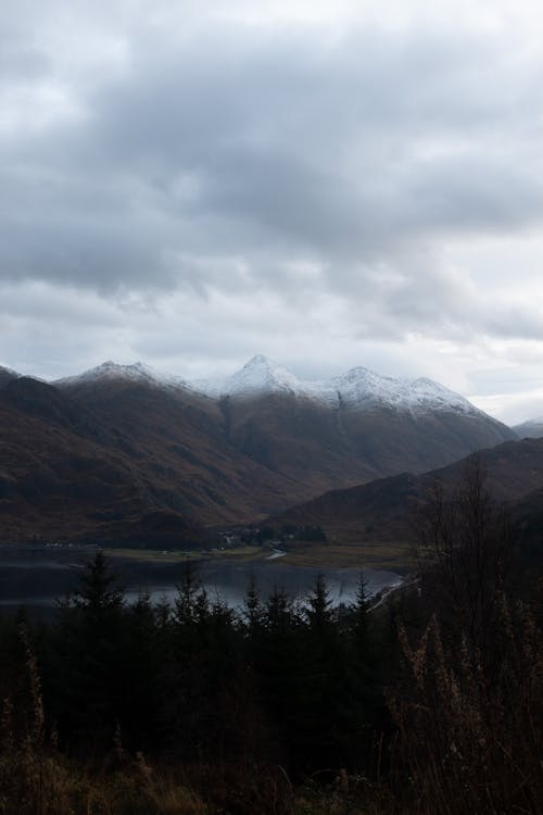 Calm lake surrounded by green coniferous trees and rough mountains with snowy peaks against cloudy sky