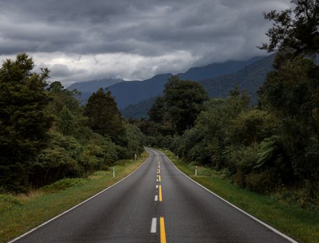 Empty asphalt road going through green trees and shrubs with hills covered with forest under gray sky