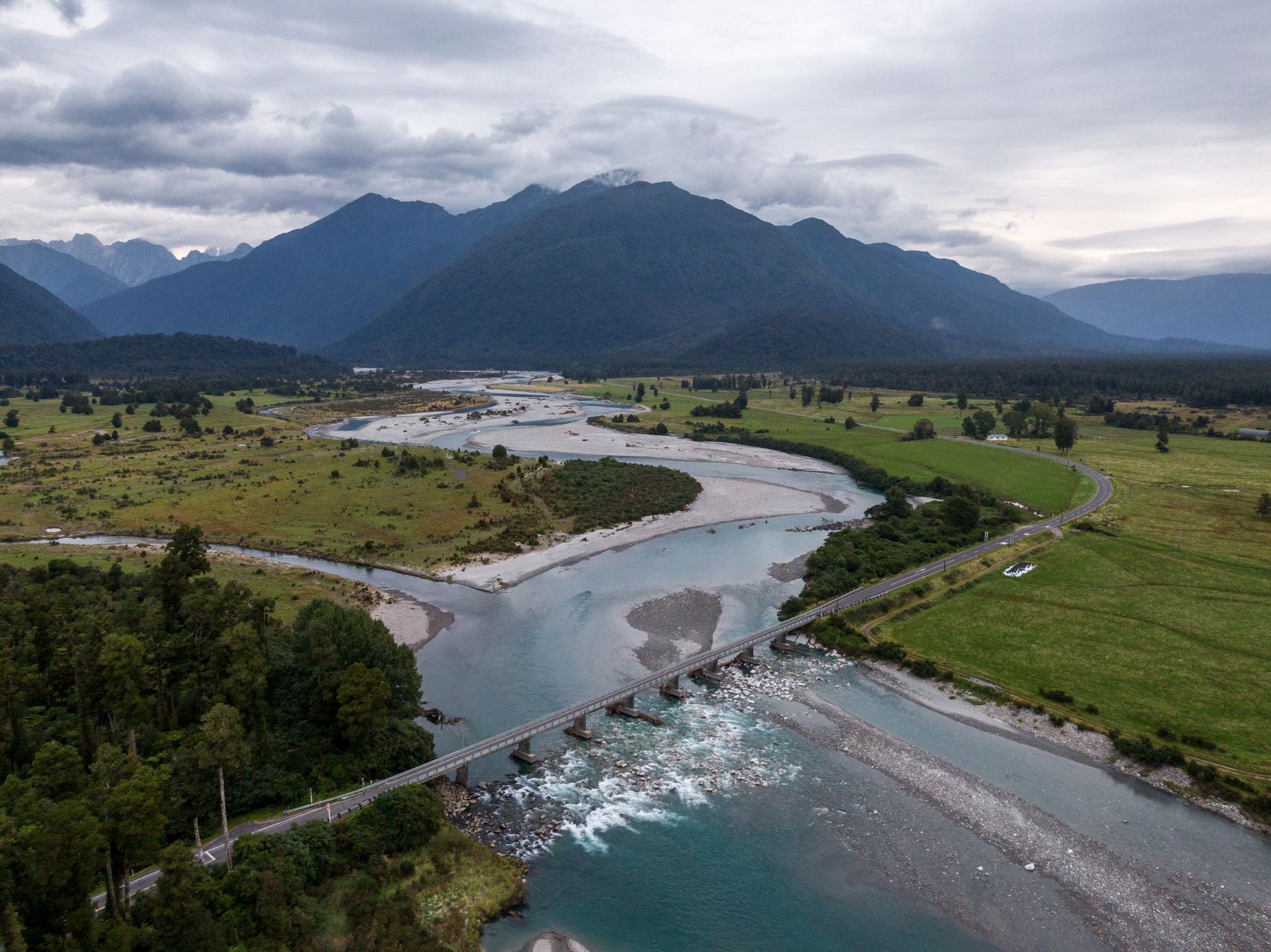 Drone view of green valley with bridge crossing meandering river against mountain covered with clouds