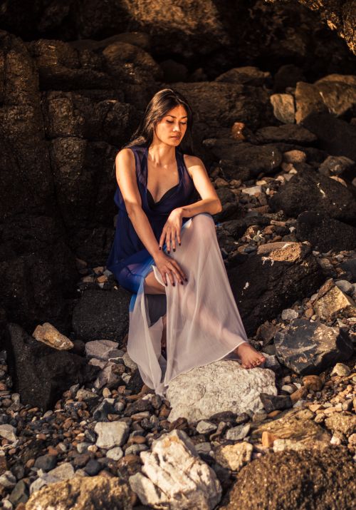 Free Elegant woman in a flowing dress poses thoughtfully on a rocky beach. Stock Photo