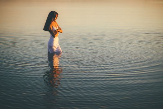 日落时分，一位身着白色连衣裙的女子静静伫立在宁静的水边