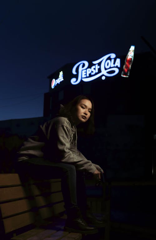 Free Full body Asian female in casual outfit sitting on bench against glowing signboard on street at night Stock Photo