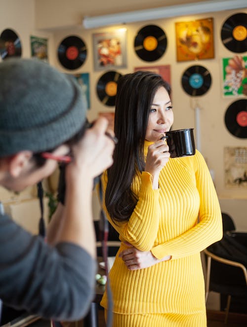 Faceless male photographer taking picture of young slim Asian woman in casual clothing drinking coffee in room decorated with paintings and vinyl disks