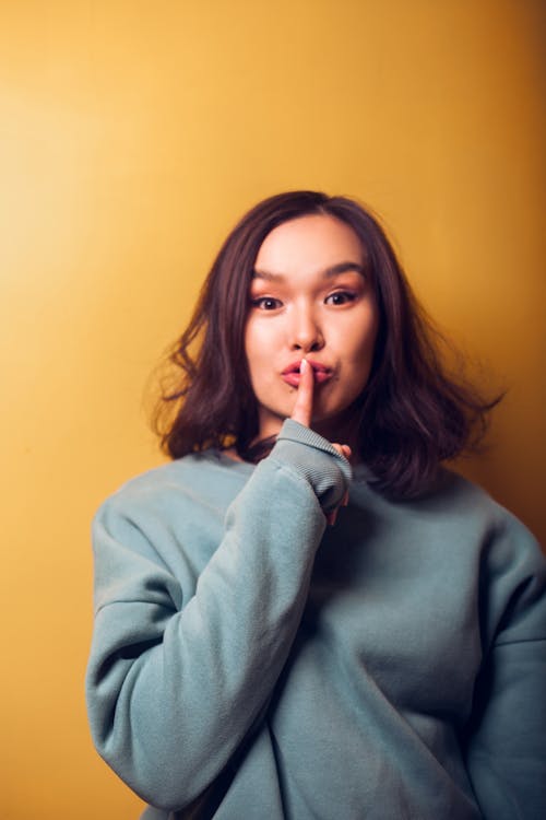 Young Asian female in warm clothing doing shh gesture with finger on lips and looking at camera while standing near orange wall