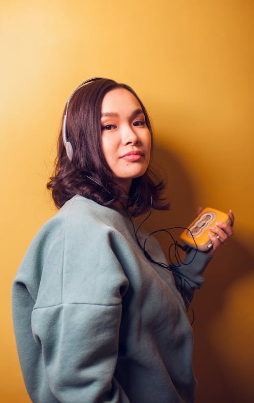 Side view of young Asian woman in warm sweater with headphones listening to music via vintage player looking at camera