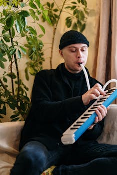 A man playing a melodion instrument indoors, surrounded by green plants, creating a cozy atmosphere.