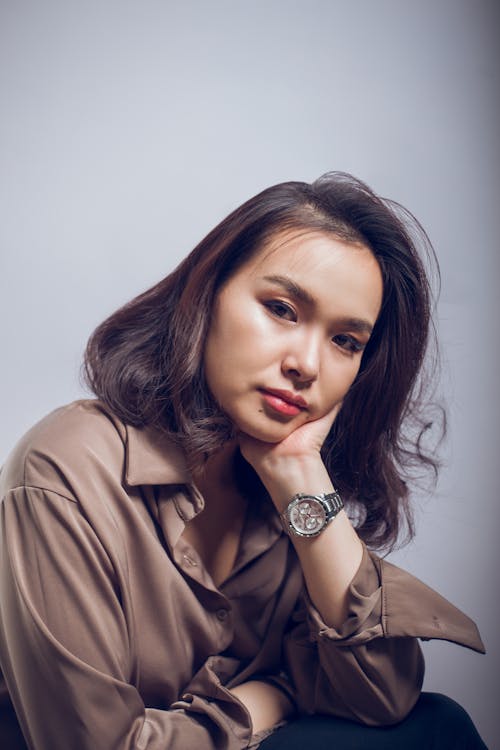 Pensive young Asian female in formal shirt with wristwatch sitting with hand under chin and looking at camera