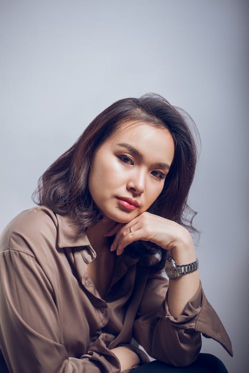 Melancholic young female employee in formal wear looking at camera while leaning to hand on gray background