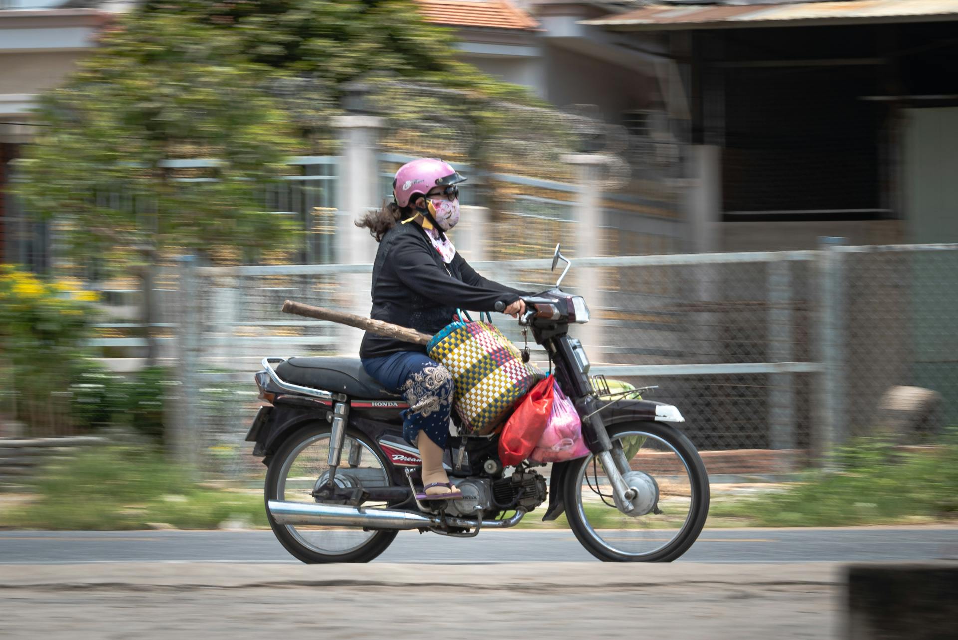 A woman wearing a helmet and face mask rides a motorcycle transporting goods on a city street.
