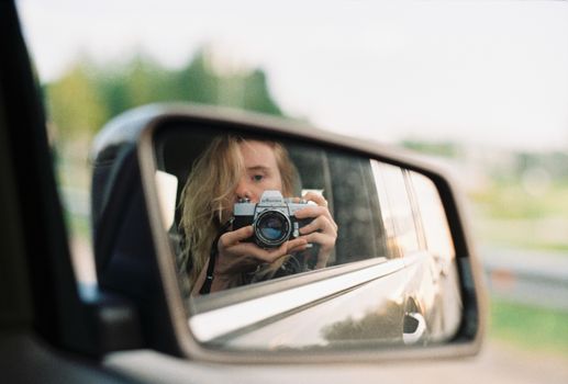 Blonde woman captures herself with an analog camera in a car's side mirror reflection.