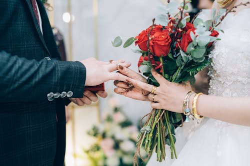 Crop bride in white dress with red flower bouquet with bracelets on wrist putting wedding ring on grooms finger