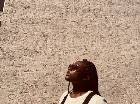 Black woman with braided hair and sunglasses looking towards the sun. Modern portrait with beige textured background.