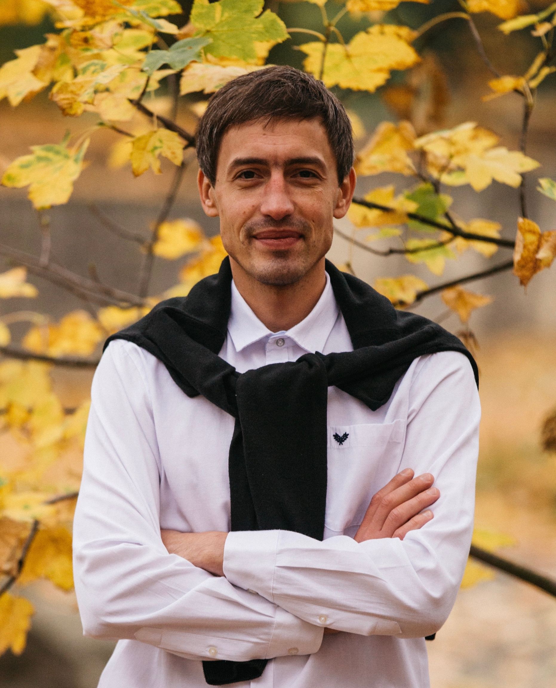 Satisfied adult ethnic male in white t shirt and black sweater on shoulders looking at camera while standing with folded arms behind colorful tree branch with maple leaves in autumn