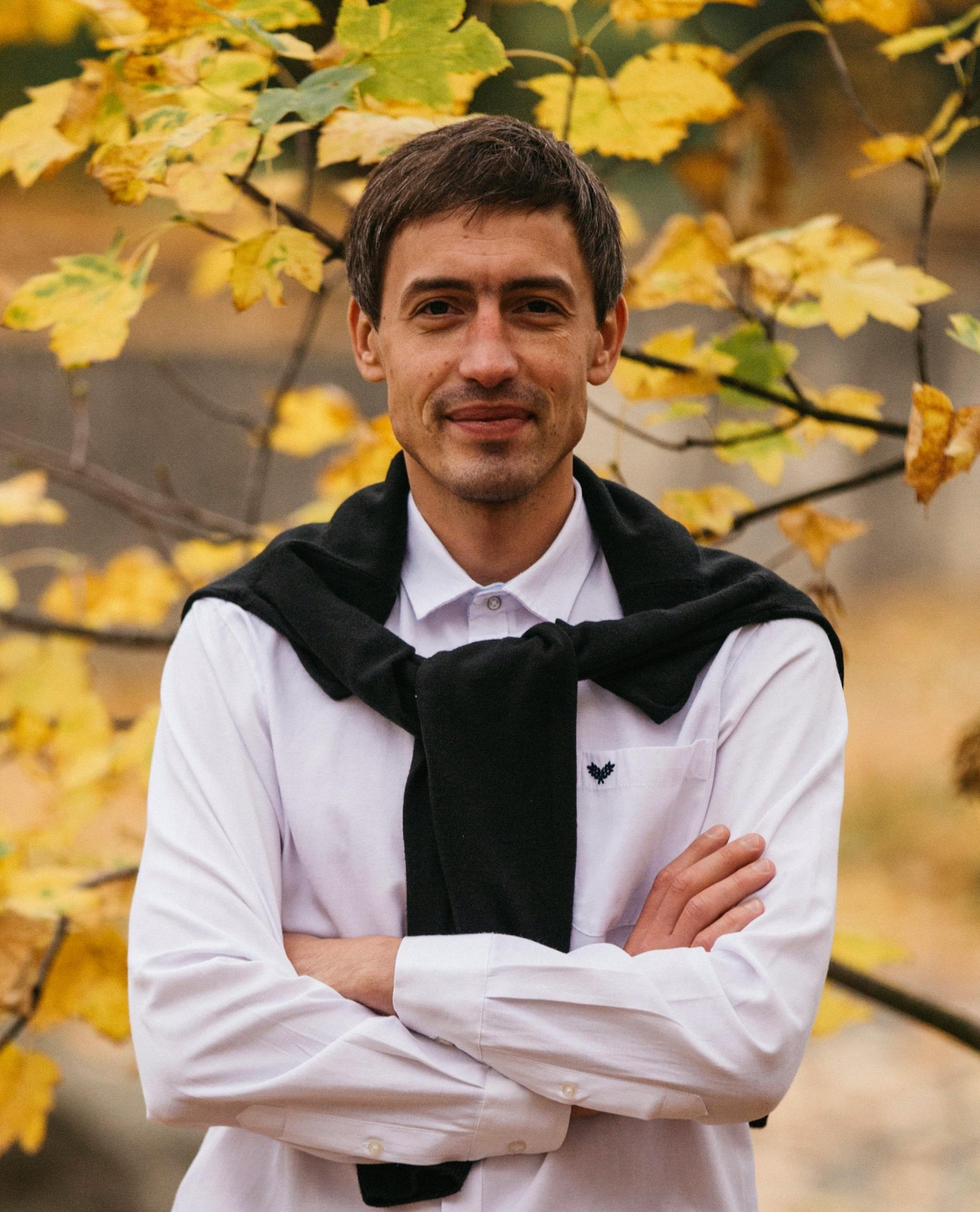Confident ethnic man standing with crossed arms behind bright tree