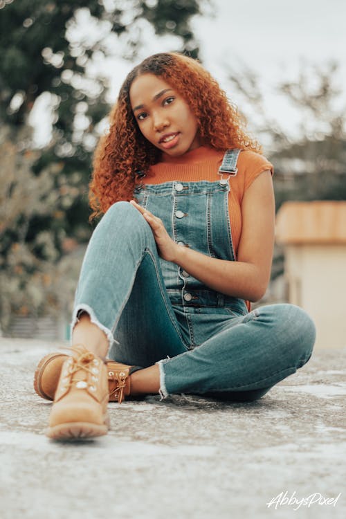 Ground level of young slim dreamy African American female with red curly hair in denim overalls sitting with tilted head on shabby walkway and looking at camera in countryside