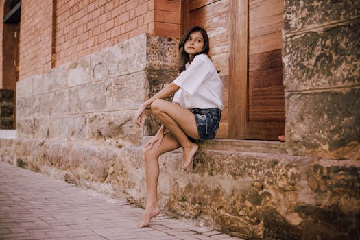 Side view of stylish slim dreamy barefoot ethnic female looking away while sitting near aged house with rough wall and wooden shutters near pavement