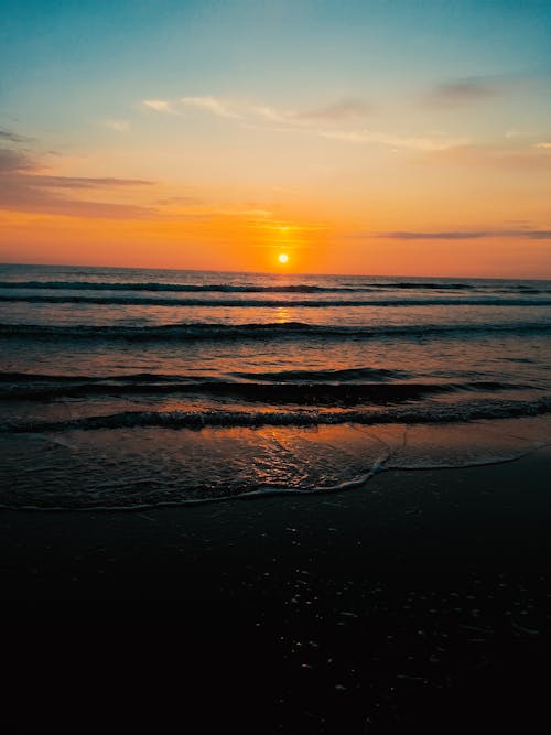 Picturesque view of sun setting over wavy foamy sea washing sandy beach at sundown