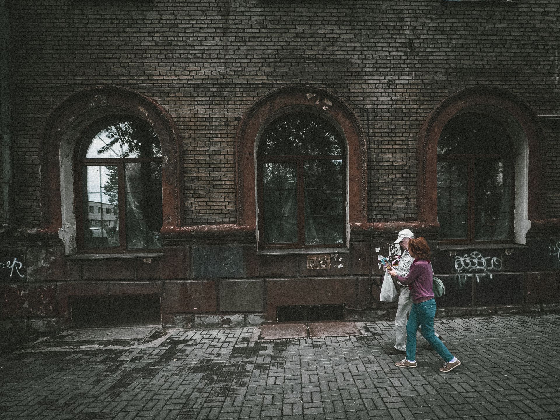 From above side view of young woman helping senior grandparent with plastic bag strolling on tiled walkway near aged masonry building in town