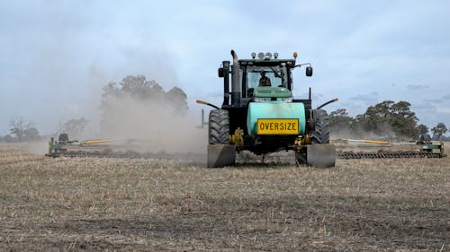 Tractor plowing agricultural field near green trees located under blue sky in summer day