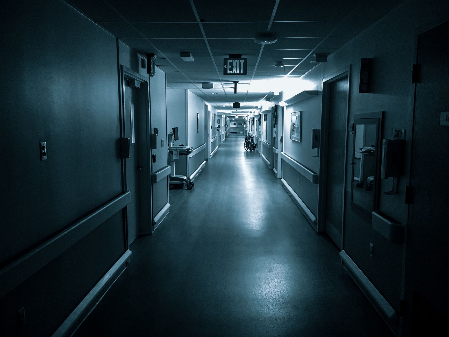 Black and white poorly lit empty wide corridor of hospital with doors on both sides and exit sign on ceiling