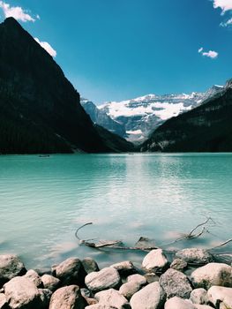 Picturesque scenery of calm lake with stones on coast located between mountains covered with snow against blue sky on sunny day