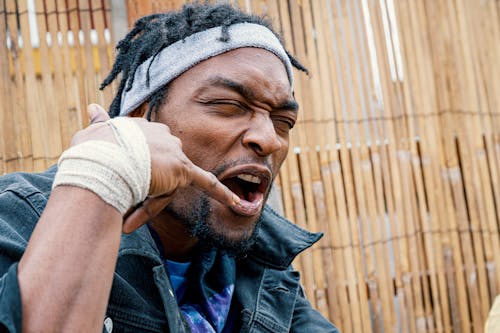 Free Frowning eccentric African American male in casual wear and headband showing call me sign with hand with bandage on wrist while spending time outside Stock Photo