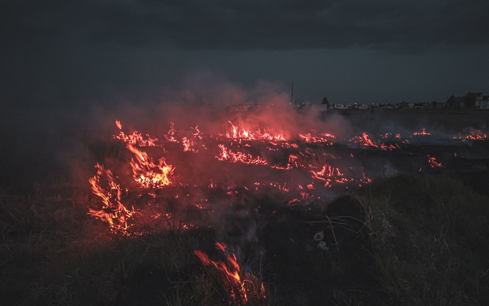 Flames and smoke moving across a dark rural field under an overcast sky.