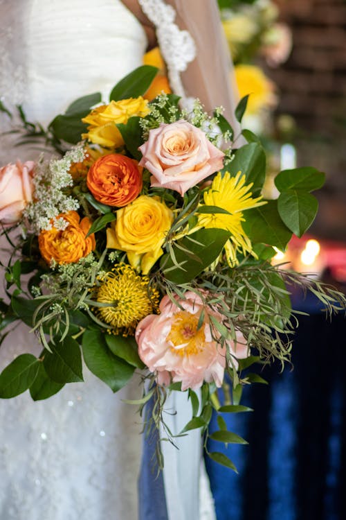 Anonymous woman in white embroidered dress holding bouquet with yellow and pink roses in greenery