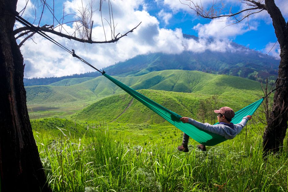 Person relaxing in a hammock amidst lush green hills under a vibrant blue sky.