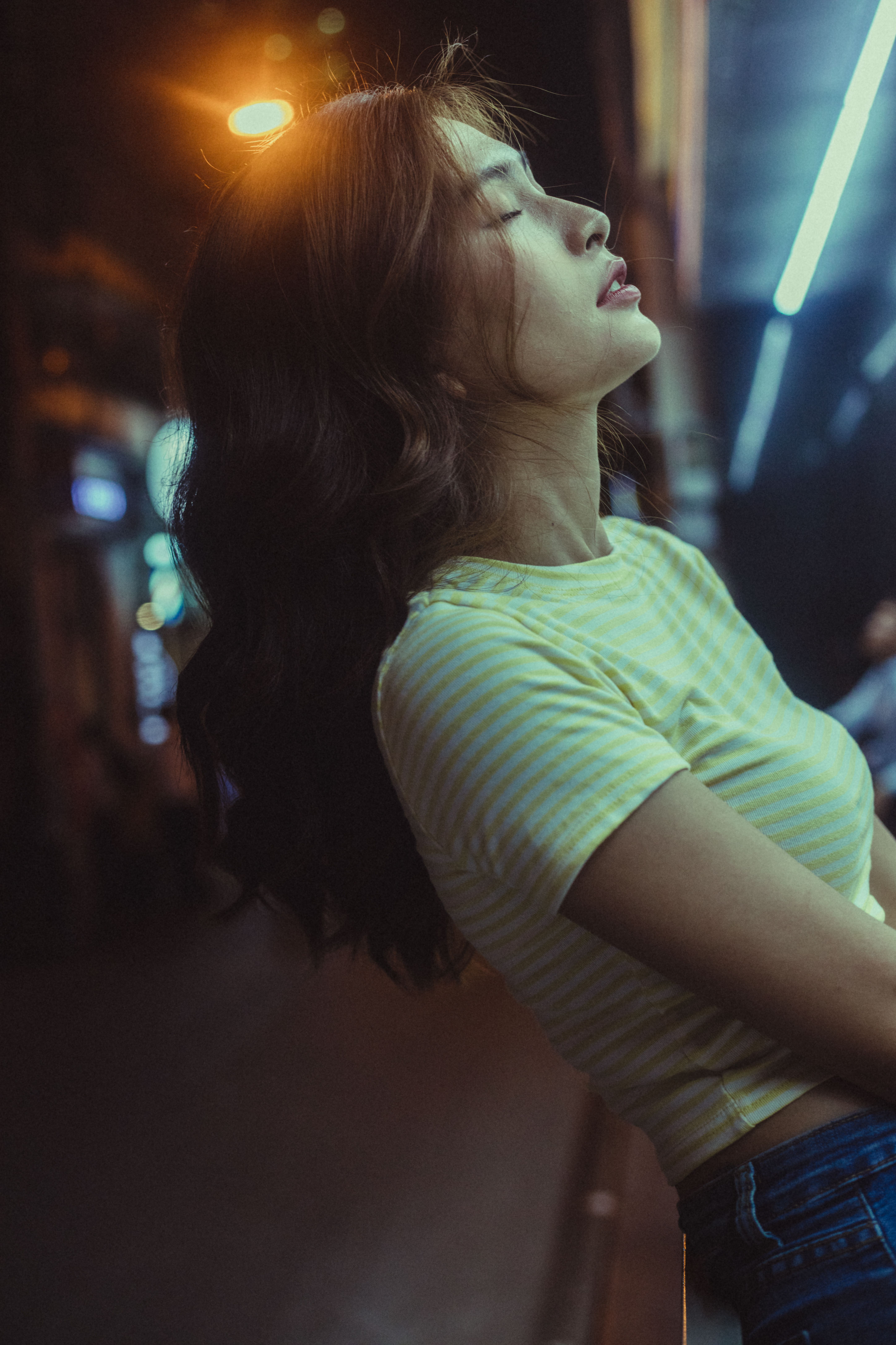 Side view of young Asian female in casual clothes leaning on railing of balcony with closed eyes against blurred urban background at night