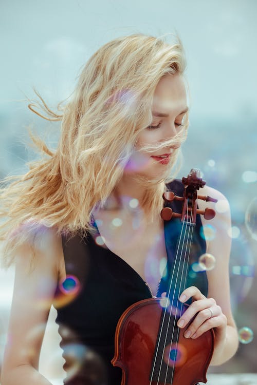 Peaceful young female musician with blond windy hair standing on street with violin among colorful soap bubbles and enjoying sunny day