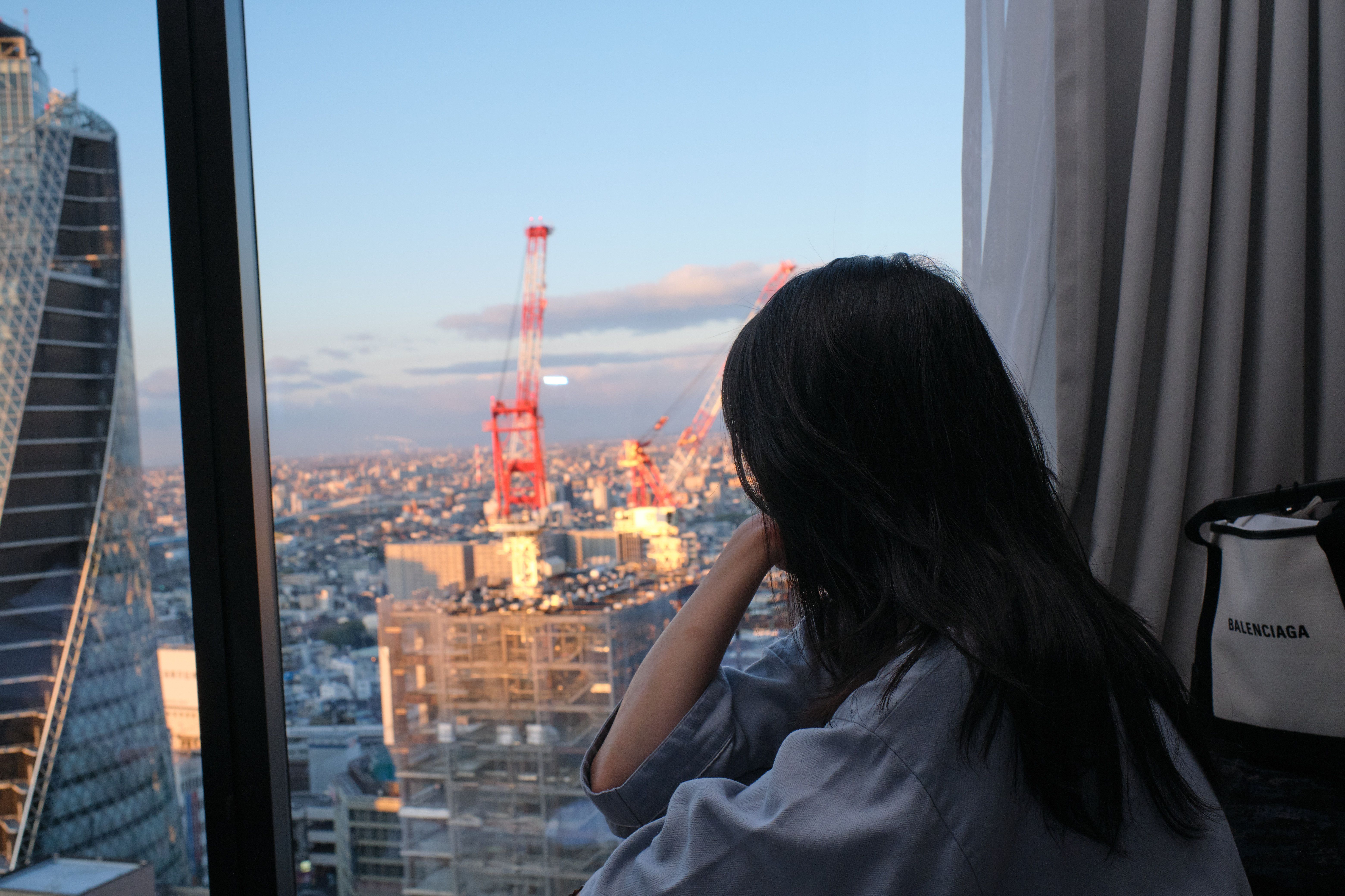 一名女子从窗户凝视着蓝天下的城市天际线，天际线上矗立着起重机