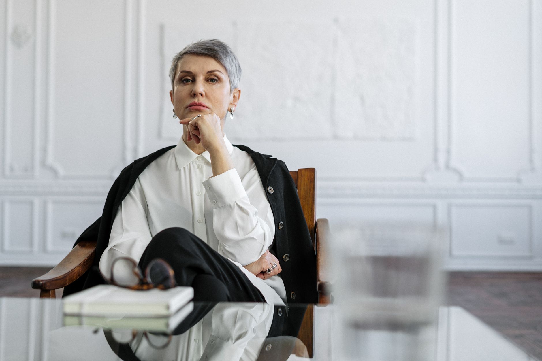 A thoughtful professional woman sits at a glass table in a well-designed office, conveying contemplation and expertise.